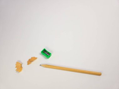 Ahşap bir kalem, yeşil bir kalemtıraş ve temiz beyaz bir yüzey üzerinde ahşap talaşı içeren basit bir natürmort kompozisyonu. Görüntü, göz seviyesindeki bir perspektiften, yumuşak ışıklandırma ve objelerin detaylarına odaklanma ile çekilir. Minima.