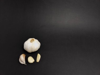 Sarımsaklı bir ampulün yakın plan çekimi ve koyu siyah arka planda üç ayrı karanfil. Sarımsak beyaz ve taze görünüyor, karanfiller pürüzsüz beyaz dokusunu gösteriyor. Kompozisyon basit, sarımsağı vurguluyor.