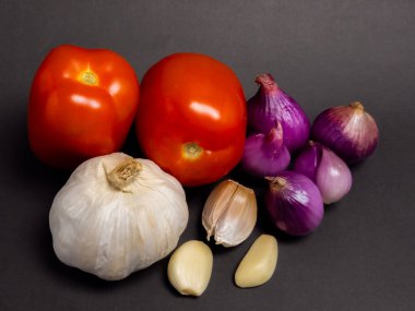 Bir natürmort fotoğrafında iki olgun kırmızı domates, bir bütün sarımsak ampul, bir sarımsak karanfil ve birkaç küçük kırmızı soğan bulunur. Bu taze sebzeler koyu gri bir arka planda sanatsal olarak düzenlenerek görsel olarak çekici bir kompozisyon oluşturulmuştur. Işıklandırma