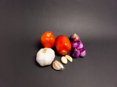 Yemek pişirmek için taze malzemeler içeren durgun bir yaşam görüntüsü. İki olgun kırmızı domates, bir bütün sarımsak ampulü, iki diş sarımsak ve koyu gri bir zemin üzerine serpiştirilmiş arpacık soğanı. Kompozisyon basit, doğal renkleri vurguluyor.