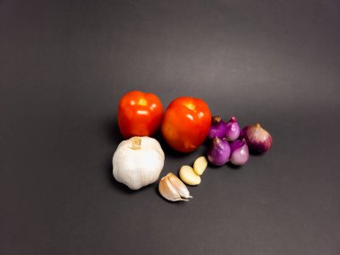 Yakın çekim bir stüdyo çekiminde katı siyah arka planda taze, pişirilmemiş sebzelerden oluşan bir koleksiyon yer alır. Kompozisyonda iki canlı kırmızı domates, birkaç kırmızı soğan, bir soğan soğanı, bir sarımsak ampulü ve birkaç karanfil yer alıyor.