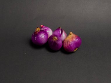 Bu yakın çekim stüdyo çekiminde koyu gri bir yüzeye dizilmiş taze, kırmızı soğan kümesi yer alıyor. Soğanlar canlı, koyu mor renkleri var, desenli derileri ve soğanlı şekilleri var. Işıklandırma onların doğal renklerini ve biçimlerini vurguluyor.