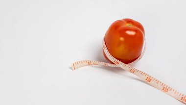 Canlı bir kırmızı domates, sağlık, diyet ve ağırlık yönetimi kavramlarını vurgulayan bir mezura ile çevrilidir. Resim temiz beyaz bir arkaplanda, özneyi vurguluyor. Kompozisyon basit ama etkili, dağınıklığı iletiyor.