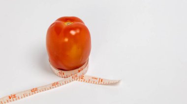 Canlı bir kırmızı domates, ağırlık yönetimi, porsiyon kontrolü ve sağlıklı yeme alışkanlıklarını vurgulayan bir mezura ile çevrilidir. Görüntü, domatese ve diyetle olan bağlantısına odaklanarak temiz beyaz bir arkaplan üzerine kurulur.