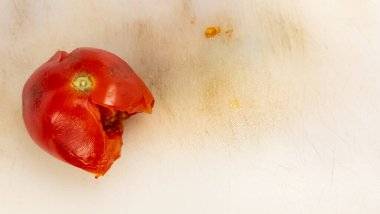Canlı kırmızı bir domatesin yakın plan çekimi, kısmen yenmiş, beyaz bir kesme tahtasının üzerinde dinleniyor. Domates, görünür tohumlar ve etle biraz düzensiz bir şekil sergiliyor. Kesme tahtası, hafif çizikler ve lekelerle kullanıldığını gösteriyor. 