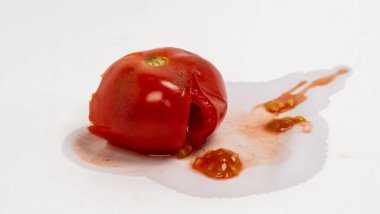 Beyaz bir yüzeyde hasarlı bir domatesin yakın plan çekimi. Kırmızı domatesin büyük bir deliği var ve ezilmiş ya da hasar görmüş gibi görünüyor. Domates suyu ve tohumlar etrafa saçılmış. Çürüme durumunu vurguluyorlar. Görüntü iyi aydınlatılmış, odaklanılmış.