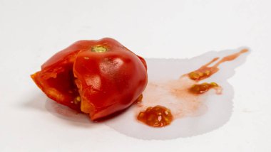 Hasarlı kırmızı domatesin yakın plan çekimi, kısmen ezilmiş, meyve suyu ve tohumlar beyaz arka plana saçılmış. Görüntü olgun meyvenin dokusunu ve rengini vurgular. Kompozisyon basittir, T 'nin tazeliğini ve doğal durumunu vurgular.