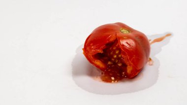 Beyaz bir yüzey üzerinde parçalanmış, olgun, kırmızı bir domatesin yakın plan çekimi. Domates kırılarak açılmış, tohumları ve suyu dökülmüş. Işıklandırma parlak ve arka plan temiz. Hasarlı meyvenin dokusunu ve detaylarını vurguluyor. İma.