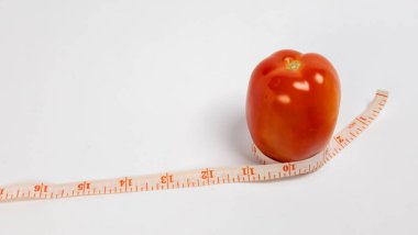 Canlı bir stüdyo çekiminde, etrafında esnek bir mezura bulunan taze, olgun bir domates görülüyor. Domates koyu kırmızı renktedir ve pürüzsüz ve sulu görünür. Şerit ölçü bej ve inç işaretli. Görüntü beyaz bir arka plana karşı iyi aydınlatılmış.