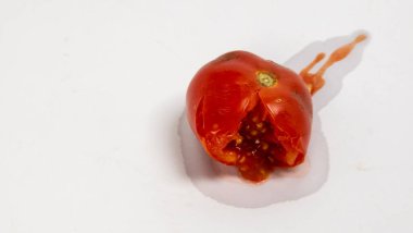 Yakından çekilen bir fotoğraf olgunlaşmış kırmızı bir domates yakalıyor ve içini ve tohumlarını ortaya çıkarıyor. Domates suyu dışarı doğru yayılmış, düz beyaz bir yüzeyde bulunur. Görüntü domatesin dokusunun dokusunu ve rengini vurguluyor.
