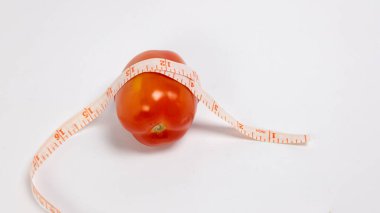 Canlı bir kırmızı domates, temiz beyaz arkaplana karşı bir mezura ile çevrilidir. Bu görüntü sağlık, diyet, kilo kaybı ve sağlıklı yeme alışkanlıklarını sembolize eder. Bileşim basit ve temiz, porsiyon boyutunu ölçme kavramını vurguluyor