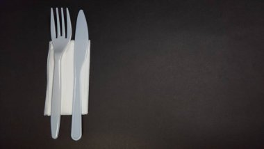 Yakın plan bir stüdyo çekiminde beyaz bir peçeteye düzgün yerleştirilmiş tek kullanımlık çatal ve bıçak kullanılıyor. Çatal bıçaklar açık gri renktedir, koyu renkli arka planla tezat oluştururlar. Kompozisyon basit, çatal bıçak ve peçete yerleştirilmiş.