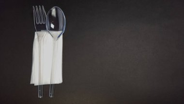 Yakın plan çekimde temiz plastikten yapılmış bir çatal ve kaşık, temiz beyaz bir peçeteye sarılmış. Çatal bıçak takımı karanlık, sade bir arka plana karşı yerleştirilmiş ve minimalist ve temiz bir kompozisyon oluşturuyor. Işıklandırma yumuşak ve eşit, vurgulu.