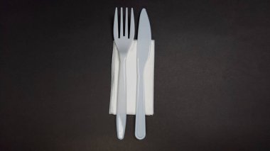 Katlanmış beyaz bir peçetenin yanına yerleştirilmiş plastik çatal ve bıçağın yukarıdan aşağı bir görüntüsü, hepsi de siyah arka planda. Bu minimalist kompozisyon nesnelerin basitliğini, dokularını ve açık renkli nesnelerin arasındaki kontrastı vurgular. 