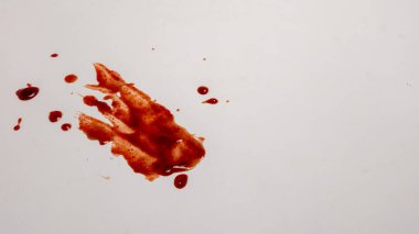 Beyaz bir yüzeye sıçramış kan lekeleri, bir suç mahalli ya da yaralanma olduğunu gösteriyor. Görüntü çeşitli damlacıklar ve daha büyük bir kan lekesi gösteriyor, dokuyu ve rengi vurguluyor. Bileşim, kanın detaylarına ve görsel etkisine odaklanıyor., 