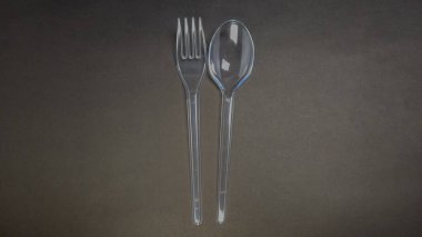 Koyu gri, desenli bir yüzey üzerinde temiz bir plastik çatal ve kaşığın yakın plan çekimi. Çatal bıçaklar dikey olarak, yan yana, basit ama görsel olarak çekici bir kompozisyon oluşturuyor. Işıklandırma plastiğin şeffaflığını vurguluyor.