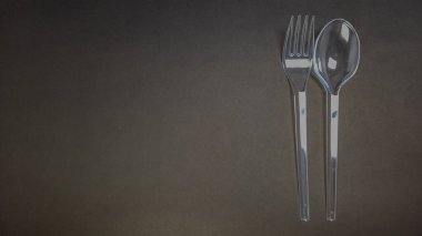 Plastik bir çatal ve kaşığın karanlık ve dokulu bir arka planda çekilmiş bir stüdyo çekimi. Çatallar çatalın sağındaki kaşıkla dikey olarak yerleştirilmiş. Görüntü basitliği, minimalizmi ve transp arasındaki karşıtlığı vurgular.
