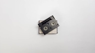 MiniDV kasetinin en alttaki görüntüsü. Kaset siyah ve çanta görünür etiketlerle şeffaf. Arka plan bembeyaz, temiz ve minimalist bir kompozisyon oluşturuyor. Görüntü yumuşak gölgelerle aydınlanmış, dokuyu vurguluyor.