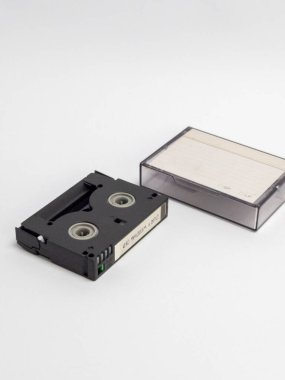 Bir MiniDV kamera bandının yakın çekimini ve beyaz arka planda izole edilmiş plastik kutusunu. Kaset siyah ve çanta şeffaf. Görüntü, bandı vurgulayan sığ bir alan derinliğiyle ışıl ışıl parlıyor. Kompozitosu.