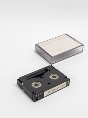 Bir fotoğrafta, temiz beyaz bir zemin üzerine kurulmuş mini bir DV kamera bandı ve temiz plastik kutusu yer alıyor. Siyah kasette gümüş makaralar ve üzerinde 
