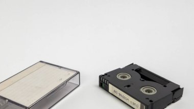 Bu görüntüde her ikisi de temiz beyaz bir yüzey üzerinde duran küçük bir DV kaseti ve koruyucu kılıfı var. Kasetlerin detaylarına ve nasıl düzenlendiklerine odaklan. Bir sürü boş alanı olan basit bir kompozisyon, onu de için uygun kılıyor.