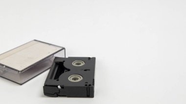 Bu görüntüde siyah bir MiniDV video kaseti ve basit, parlak beyaz bir arkaplan üzerine yerleştirilmiş şeffaf plastik kaset yer almaktadır. Kompozisyon, temiz ve modern bir estetik sunarak kasetin tasarımı ve net durumu vurguluyor. Işıklar eşit.