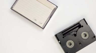 Yüksek açılı bir çekimde bir VHS kaset ve sade beyaz zemin üzerinde duran bir dava görülüyor. Siyah bant açık duruyor ve içerideki bileşenleri ortaya çıkıyor. Bu arada dosya açık ve etiketlenmek üzere çizilmiş kağıtlar sergileniyor. Kompozisyon temiz.