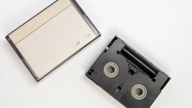 Yüksek açılı bir çekim, mini bir VHS kaseti ve kasetini düz beyaz bir yüzey üzerinde gösterir. Siyah bant açık, makaralar ortaya çıkıyor, çanta ise yakında bir yerde, etiketli. Görüntü temiz ve iyi ışıklandırılmış, çeşitli medya ve nostalji için uygun