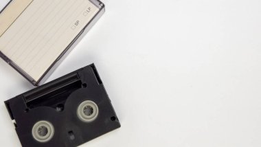 Siyah bir video kasetin ve şeffaf plastik kutusunun beyaz arka planda duran yakın plan çekimi. Dava, kayıt hızları için seçenekleri içeren bir kağıt eklemesini içeriyor ve kasetin kendisi de makaraları gösteriyor. Kompozisyon si