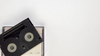 Temiz beyaz bir arkaplan üzerine kurulmuş siyah bir VHS kasetin yukarıdan aşağı görüntüsü. Kompozisyon, nostalji temalı projeler veya eski medya konseptleri için ideal olan retro teknolojiyi vurguluyor. Çoklu kopyalama alanı mevcut 