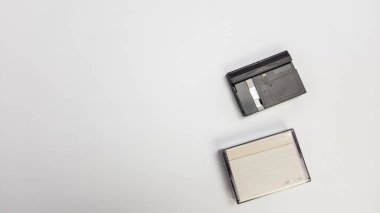 Tepeden tırnağa çekilmiş bir karede, temiz beyaz bir arkaplanda bir MiniDV kaset ve ilgili video kamera sergileniyor. Düzenleme basit ve minimalist, teknolojik bileşenleri vurguluyor. Görüntü PR ile ilgili projeler için resim uygundur