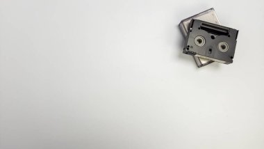 Düz beyaz arka planda iki miniDV kasetin yukarıdan aşağı görüntüsü. Siyah bant diğerinin üstünde. Görüntü, minimalist bir kompozisyonda geniş bir kopya alanı sunuyor. Odak noktası retro teknolojisi ve tasarımın basitliği..