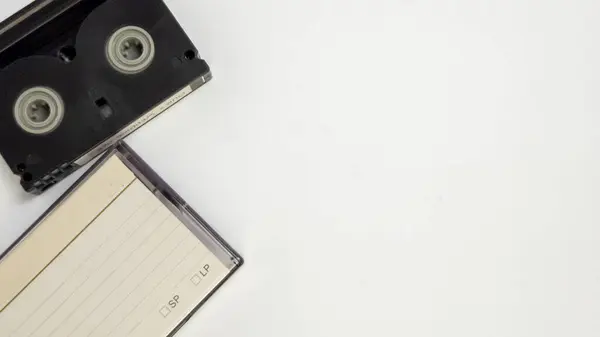 Siyah bir video kaset ve beyaz bir yüzeyde duran boş bir etiket içeren yakın plan bir çekim. Kompozisyon basit, bant ve etiket çerçevenin sol tarafını kaplıyor. Etiketin satırları ve kutucuk seçenekleri var. Görüntü...