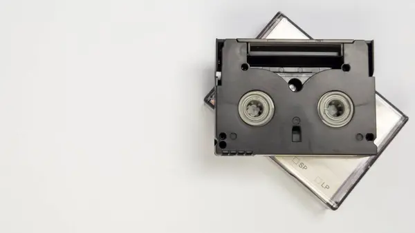 İki küçük kasetin üzerinde duran siyah bir MiniDV kaseti basit bir beyaz arka planda sergileniyor. Görüntü, kasetlerin iç bileşenleri ve etiketlemeleri de dahil olmak üzere detaylarını vurguluyor. Kompozisyonda bol miktarda kopya var.