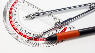 Detaylı bir stüdyo çekiminde şeffaf bir plastik iletki, metal bir pusula ve siyah turuncu bir kalem bulunur. Kompozisyon, jeomet için kullanılan araçları gösteren keskin odaklı araçları vurgular