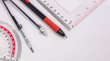 Tepemizde parlak beyaz bir zemin üzerine kurulmuş temel geometri aletleri ve okul malzemelerinden oluşan bir koleksiyon var. Kompozisyon bir iletki, pusula, cetvel ve kalem içeriyor, hepsi düzgünce düzenlenmiş. Yakın çekim odağı de vurguluyor