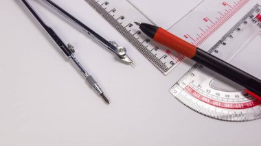 Temiz beyaz bir yüzey üzerinde düzenlenmiş pusula, cetvel, açıölçer ve kalem gibi çeşitli geometrik araçları gösteren yakın plan bir çekim. Araçlar düzgün yerleştirilmiş, detaylar ve işlevsellikleri vurgulanıyor. Kompozisyon iyi aydınlatılmış, vurgulanıyor.