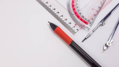 Tepeden çekimde cetvel, açıölçer ve temiz beyaz bir yüzeyde düzenlenmiş bir pusula gibi geometri araçları sergilenir. Kırmızı saplı siyah bir kalem de mevcuttur. Aletler düzgün yerleştirilmiş, hassasiyet ve doğruluğu gösteriyor.. 
