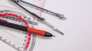 Bu resim temiz beyaz bir yüzey üzerinde düzenlenmiş temel taslak araçlarının bir koleksiyonunu gösterir. Kompozisyon bir iletki, bir pusula, bir cetvel ve bir kalem içerir. Araçlar, ayrıntılarını ve dokularını vurgulayan yakın çekim görüntülerinde sunuluyor