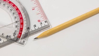 Temel okul ve ofis malzemelerini içeren yakın plan bir çekim. Şeffaf bir açıölçer, bir cetvel ve bir tahta kalem de dahil. Nesneler beyaz bir arkaplanda, temiz ve dağınık bir kompozisyon oluşturuyorlar. Odaklanma keskin, Highli.
