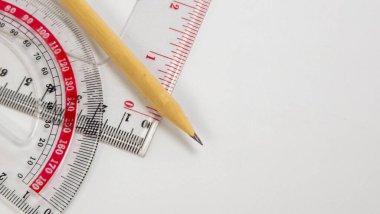 Yakın çekim bir kalem, bir açıölçer ve beyaz bir yüzey üzerinde bir cetvel içerir. Bileşim geometrik araçları vurgular, temiz ve minimalist bir estetik yaratır. Kalem ucundaki keskin odak ve ölçümün net işaretleri