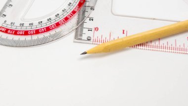 Keskinleştirilmiş bir kalemin yakın plan çekimi. Plastik bir cetvel ve açıölçer üzerinde dinleniyor. Araçlar temiz, beyaz bir yüzeye yerleştiriliyor ve geniş negatif alanı olan çekici bir bileşim oluşturuyor. Resim dr için kullanılan araçları vurgular