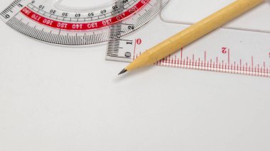 Beyaz bir yüzey üzerinde okul malzemelerini gösteren yakın plan bir çekim. Görüntü, tahta bir kalem, kırmızı işaretli şeffaf bir plastik taşıyıcı ve temiz bir plastik cetvel içerir. Kompozisyon basit, araçlara odaklanarak, bir p hissi yakalamak