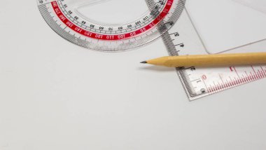Bu resim, beyaz bir arkaplanda düzenlenmiş temel kırtasiye eşyalarının yakın plan perspektifini gösterir. Kompozisyonda bir kalem, bir açıölçer ve bir küme kare, çizim, tasarım ve geometride kullanılan tüm yaygın araçlar yer almaktadır. Işıklandırma vurgusu