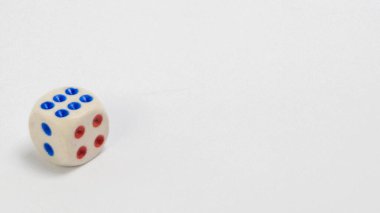 Mavi ve kırmızı noktalı beyaz bir zarın yakın plan çekimi, merkezden biraz uzakta, temiz beyaz bir arkaplanda. Işıklandırma yumuşak ve kompozisyon basit, nesneye dikkat çekiyor. Zarda iki kenar ve altı nokta var.