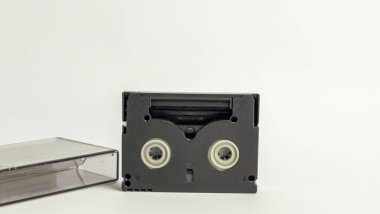 Bu resim, yanında çantası olan küçük bir DV kasetinin yakın çekimini gösteriyor. Siyah kaset ortada, şeffaf bavul da solda. Arka plan düz beyaz, temiz ve basit bir estetik sağlıyor. Bu...