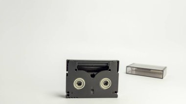 Yakın çekim bir MiniDV kaseti ve şeffaf koruyucu kılıfı beyaz arka planda yer alır. Siyah bant ortalanmış ve dava biraz bulanık. Görüntü, geçmiş videonun teknolojisini ve biçimini vurgular