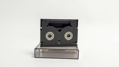 MiniDV kasetinin koruyucu kılıfının üstüne yerleştirilmiş yakın çekim bir stüdyo görüntüsü. Bant ve çantanın siyah plastiği düz beyaz arka plana karşı güçlü bir kontrast oluşturuyor. Kompozisyon basit ve temiz, fünyeyi vurguluyor.