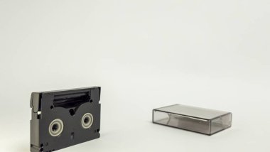 Bir MiniDV video kaseti ve temiz plastik kutusu temiz, sade beyaz bir arka planda izole edilir. Siyah bant kaseti solda, çanta da sağda duruyor. Görüntü iyi aydınlatılmış ve objelere odaklandığını gösteriyor, ideal.