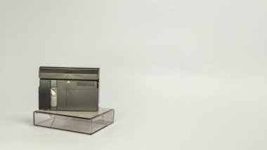 MiniDV kamerasının, beyaz bir arkaplana karşı şeffaf plastik kutusunun üzerinde dinlenirken çekilmiş yakın çekim bir fotoğrafı. Siyah kamera çantanın üzerine yerleştirilmiş, basit ve temiz bir kompozisyon sağlıyor. Görüntü retro teknolojiyi vurguluyor.